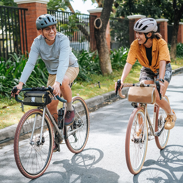 Zwei junge Erwachsene fahren lachend auf Trekking- und City-Bikes durch eine grüne Wohnstraße – Diermann - Bergsieger GmbH